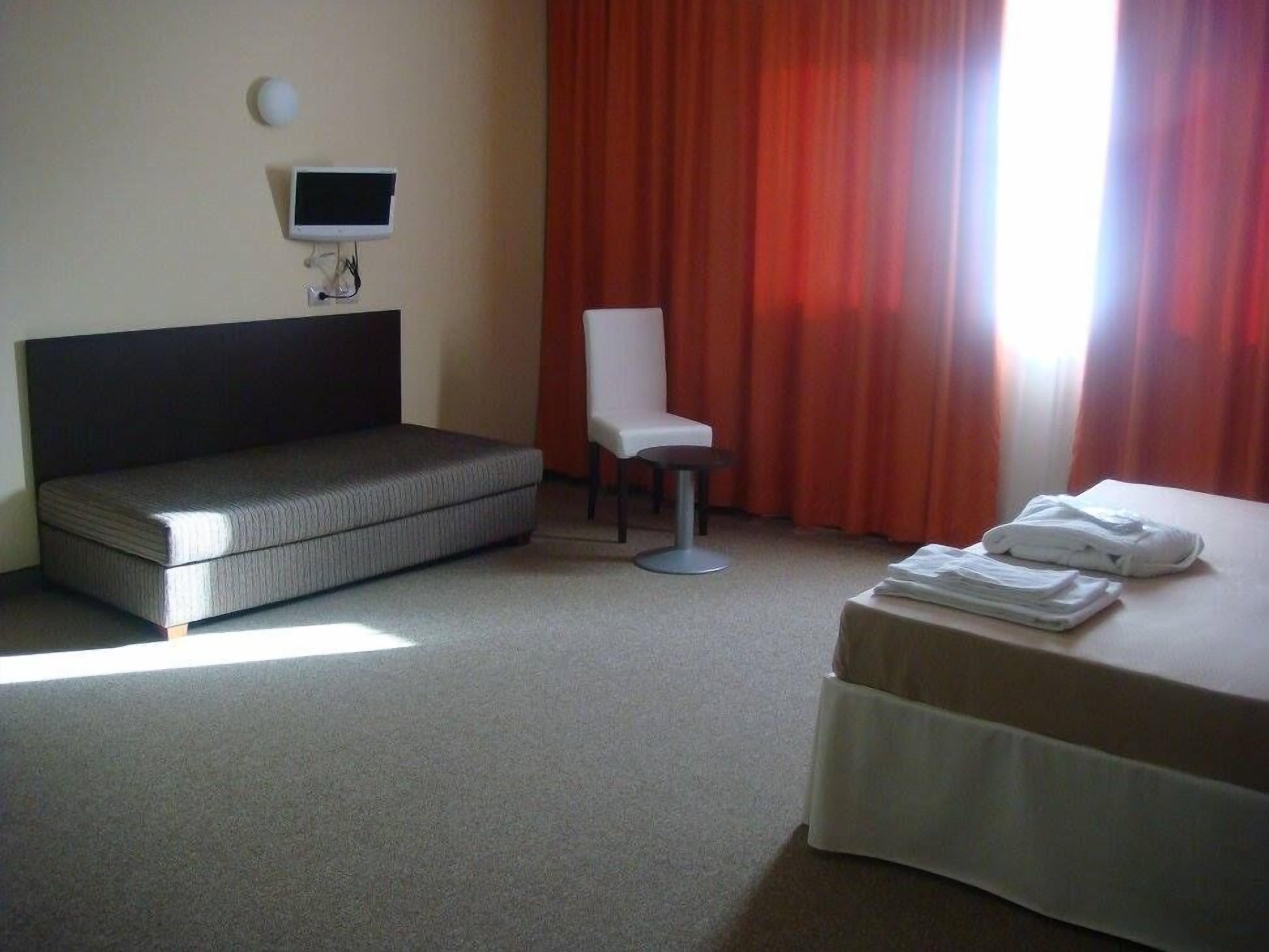 Una camera d'albergo con un letto, una sedia, un tavolino, una televisione e una finestra con tende.