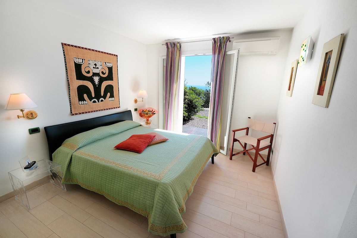 Ein Schlafzimmer mit einem Bett, einem Sessel, einem Nachttisch, einer Wandlampe und einem Fenster mit Vorhängen.