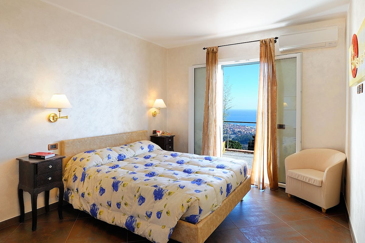 Ein Schlafzimmer mit einem Bett, zwei Nachttischen, einem Sessel und Blick auf die Stadt durch ein Fenster.