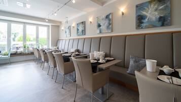 A modern dining area with a long bench seating arrangement, tables with white tableware, and abstract wall art.