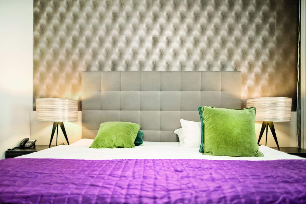 A hotel room with a tufted headboard, two green pillows, a white pillow, a purple bedspread, and two bedside lamps.