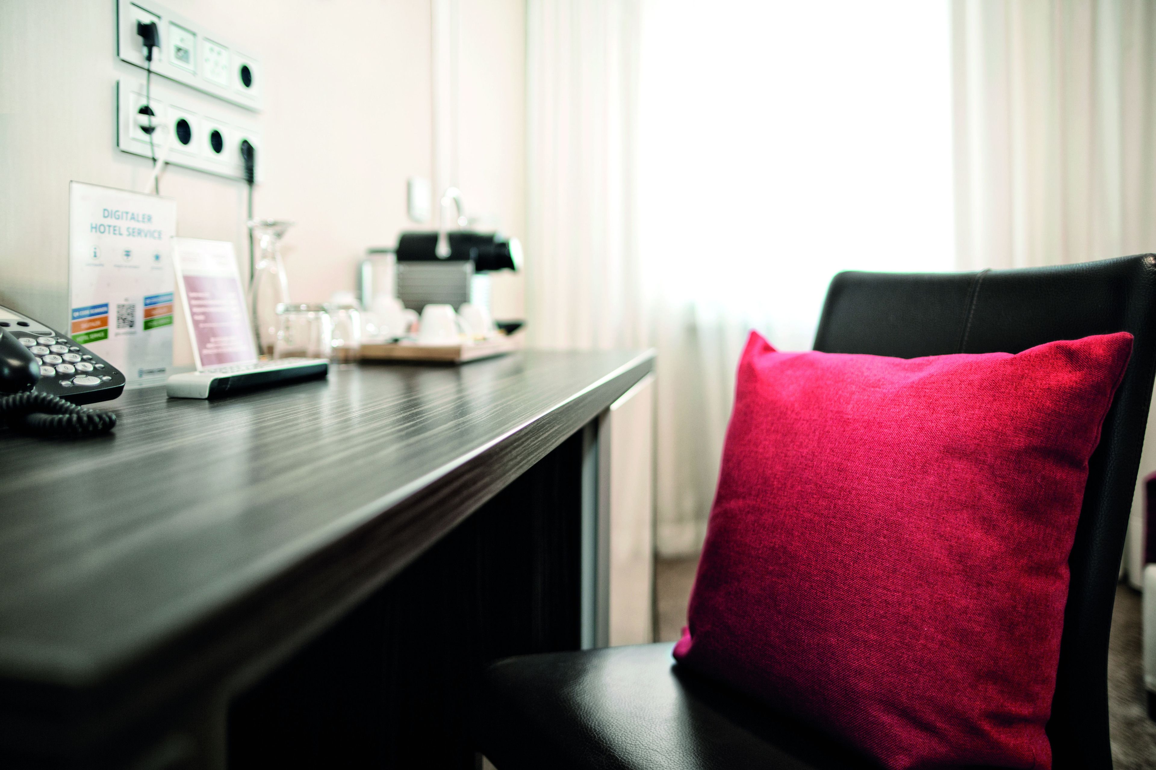 A hotel room with a desk, a telephone, a digital service sign, and a red pillow on a chair.