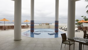 Un área de piscina en la azotea con sillones de descanso y sombrillas, rodeada de columnas y vistas al horizonte de la ciudad.