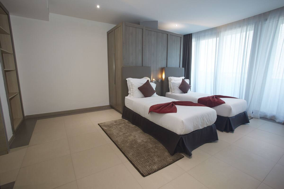 A modern hotel room with two beds, a large wardrobe, and a view of the outdoors.