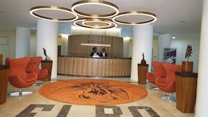 Zona de recepción de un hotel con diseño moderno, que cuenta con un mostrador central, dos recepcionistas y un gran logotipo en el suelo.