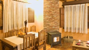 Um quarto aconchegante com uma mesa de jantar de madeira, cadeiras, uma parede de pedra, um fogão a lenha e uma janela com cortinas.
