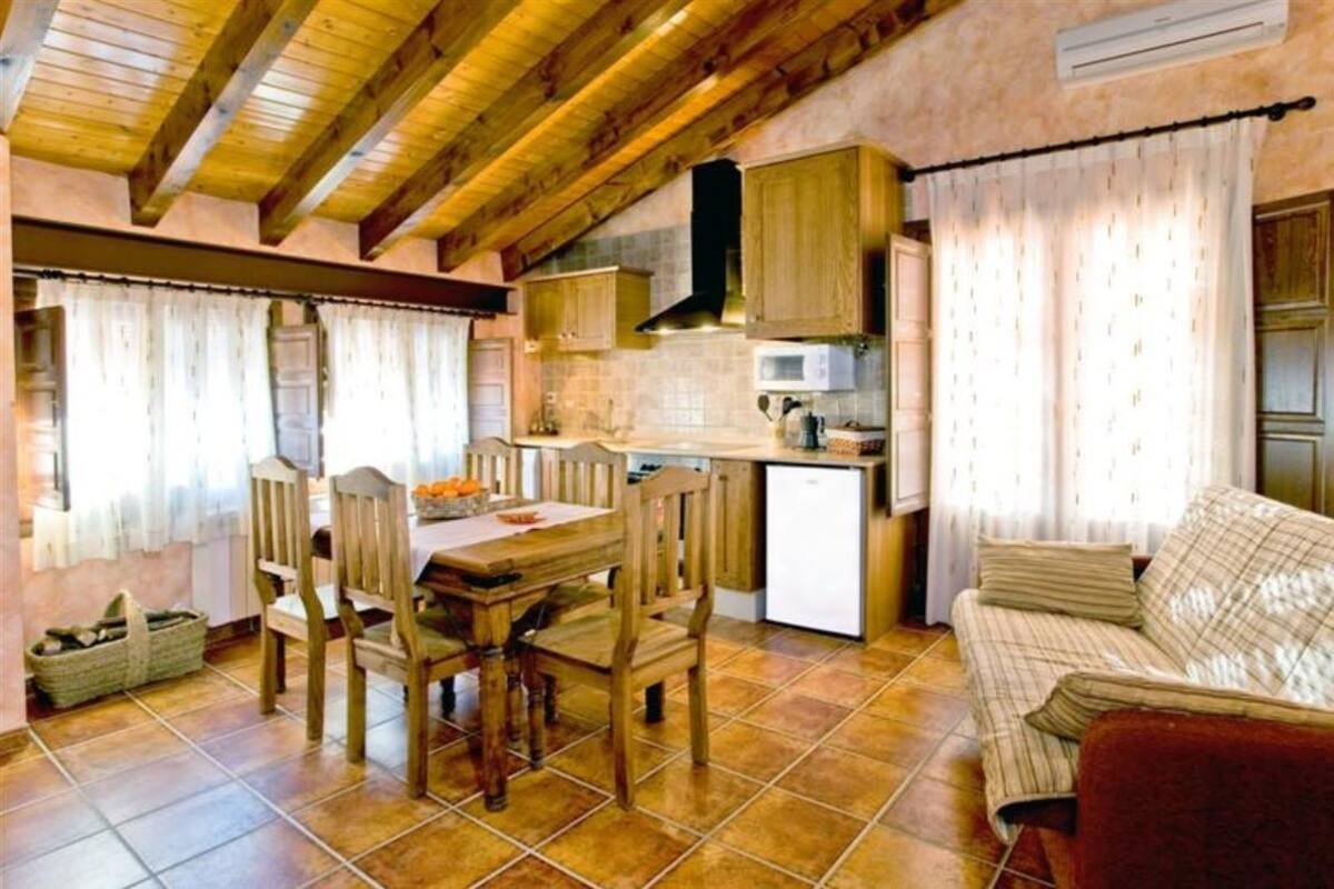 Cuina rústica amb bigues de fusta, taula de dinar amb caires, nevera petita i sofà.