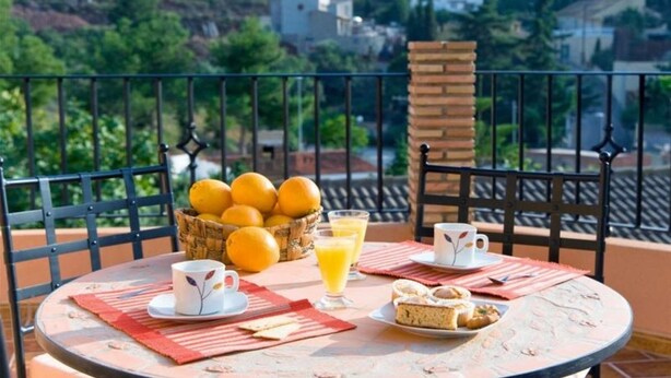 Uma mesa redonda com uma cesta de laranjas, dois copos de suco de laranja, um prato de pastelaria e uma chávena numa varanda com vista para uma encosta.
