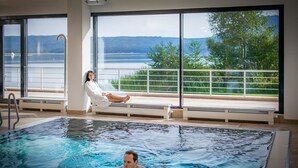 Ein Schwimmbad mit einer Person, die an einem Fenster auf einer Bank sitzt, einer anderen Person, die schwimmt, und Blick auf einen See und Berge.