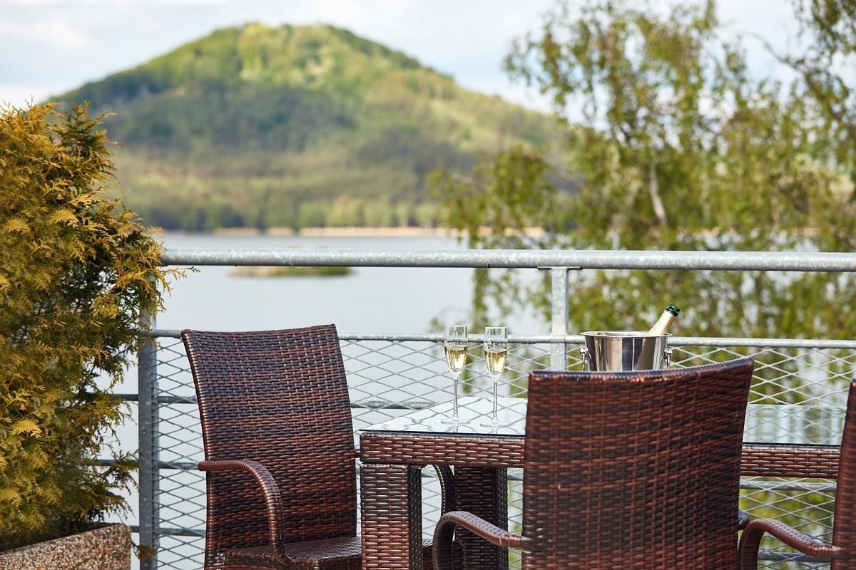 Een rieten tafel met twee stoelen en een metalen leuning, met uitzicht op een meer met een heuvel op de achtergrond.
