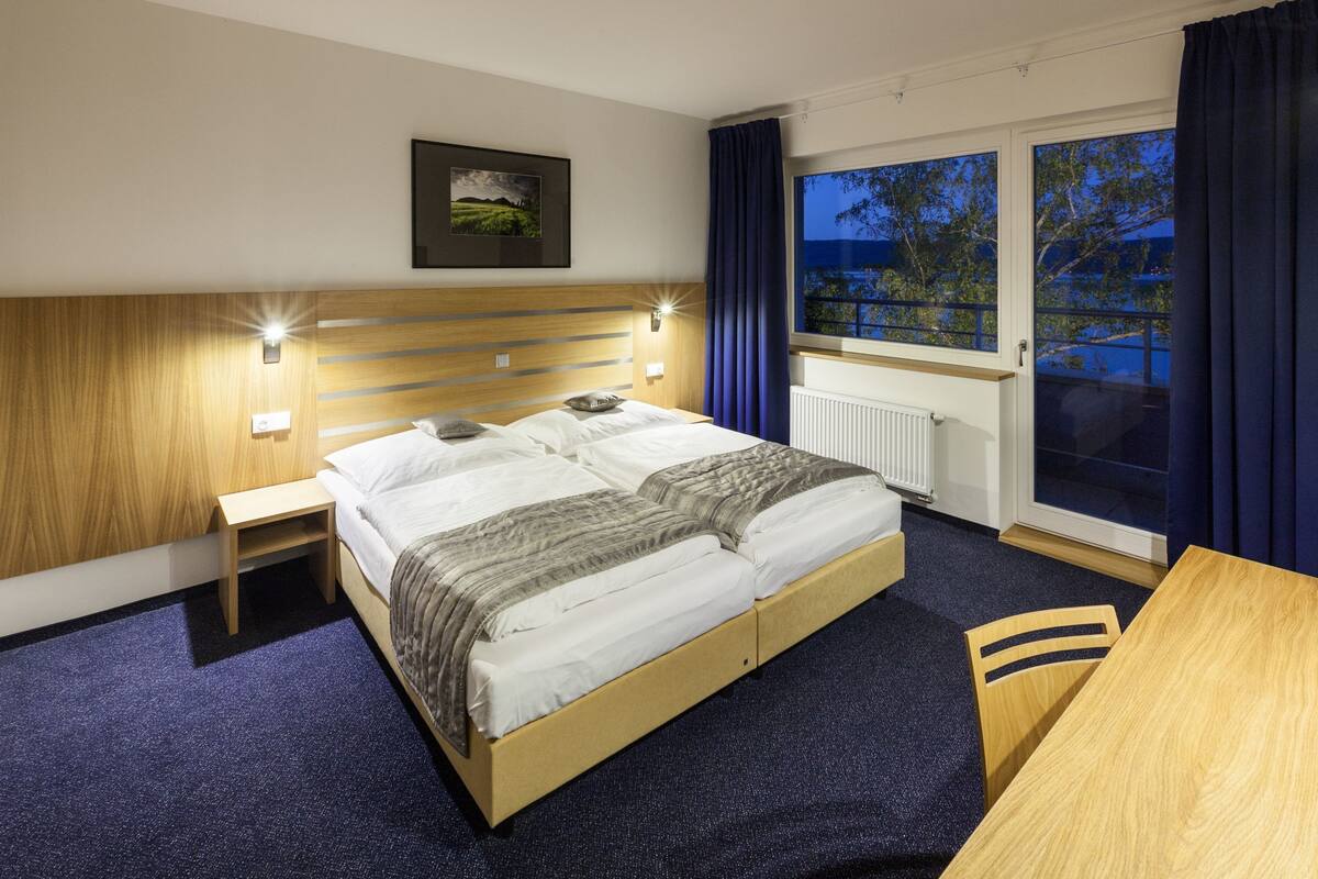 A hotel room with a double bed, bedside tables, a window with curtains, and a view of the outdoors.