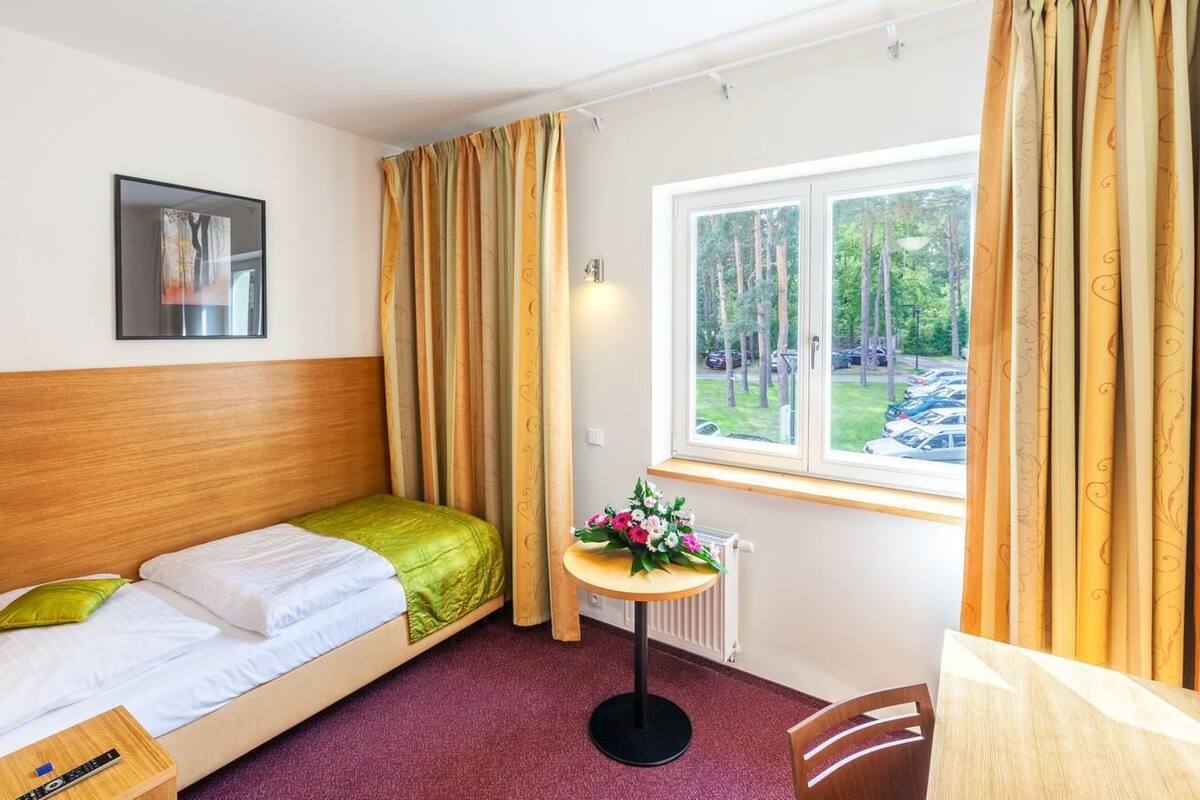 A small, single-bed room with a wooden headboard, a small table with a flower arrangement, and a window with curtains.