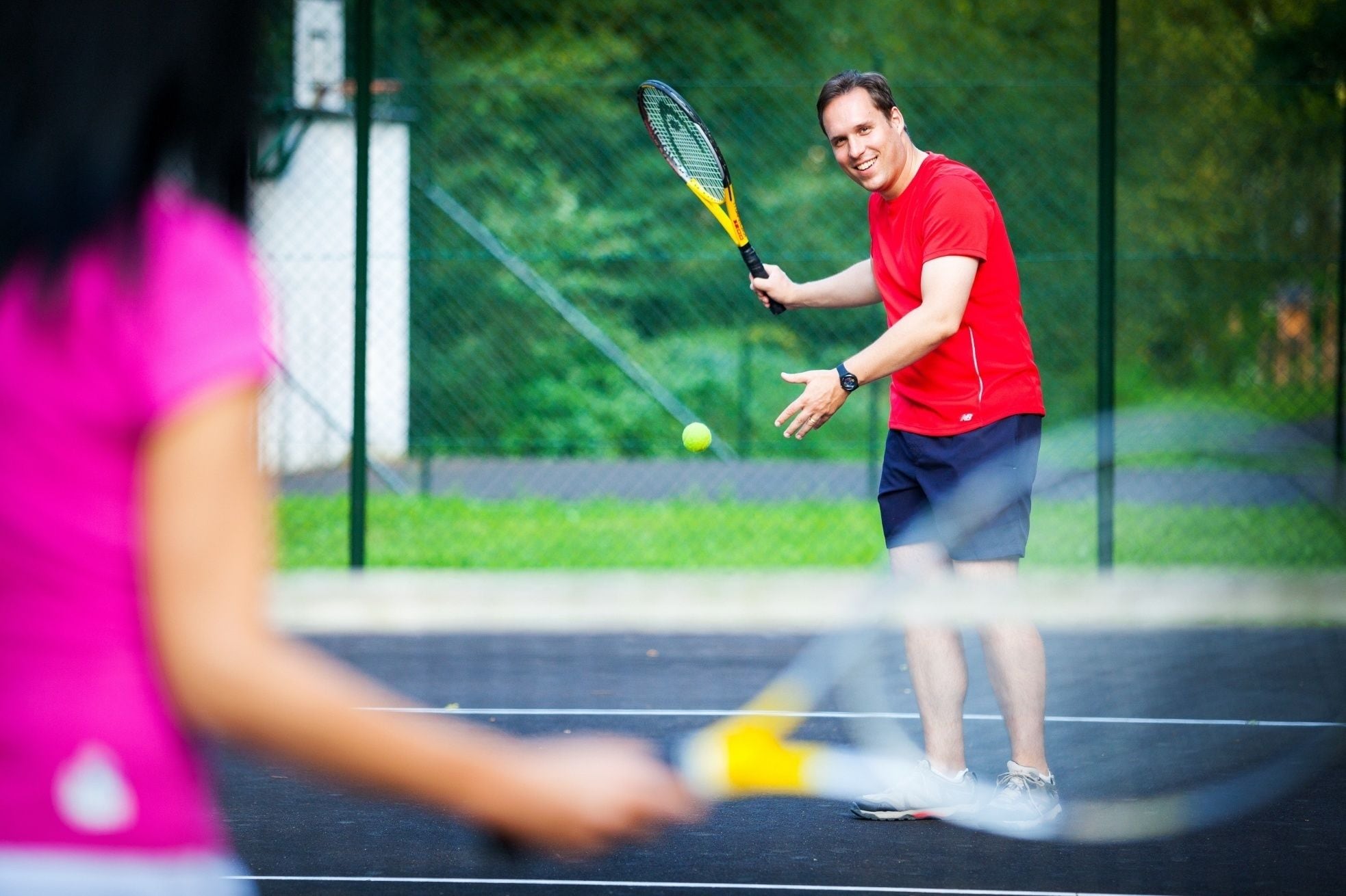 Una cancha de tenis con red, un jugador con camisa roja y una pelota de tenis en movimiento.
