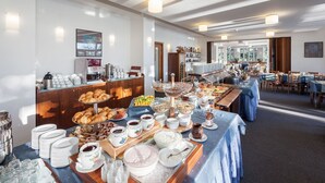 Ein reichhaltiges Buffet mit verschiedenen Speisen und Getränken, darunter Croissants, Früchte und Milchprodukte, in einem hell erleuchteten Raum mit Tischen und Stühlen.