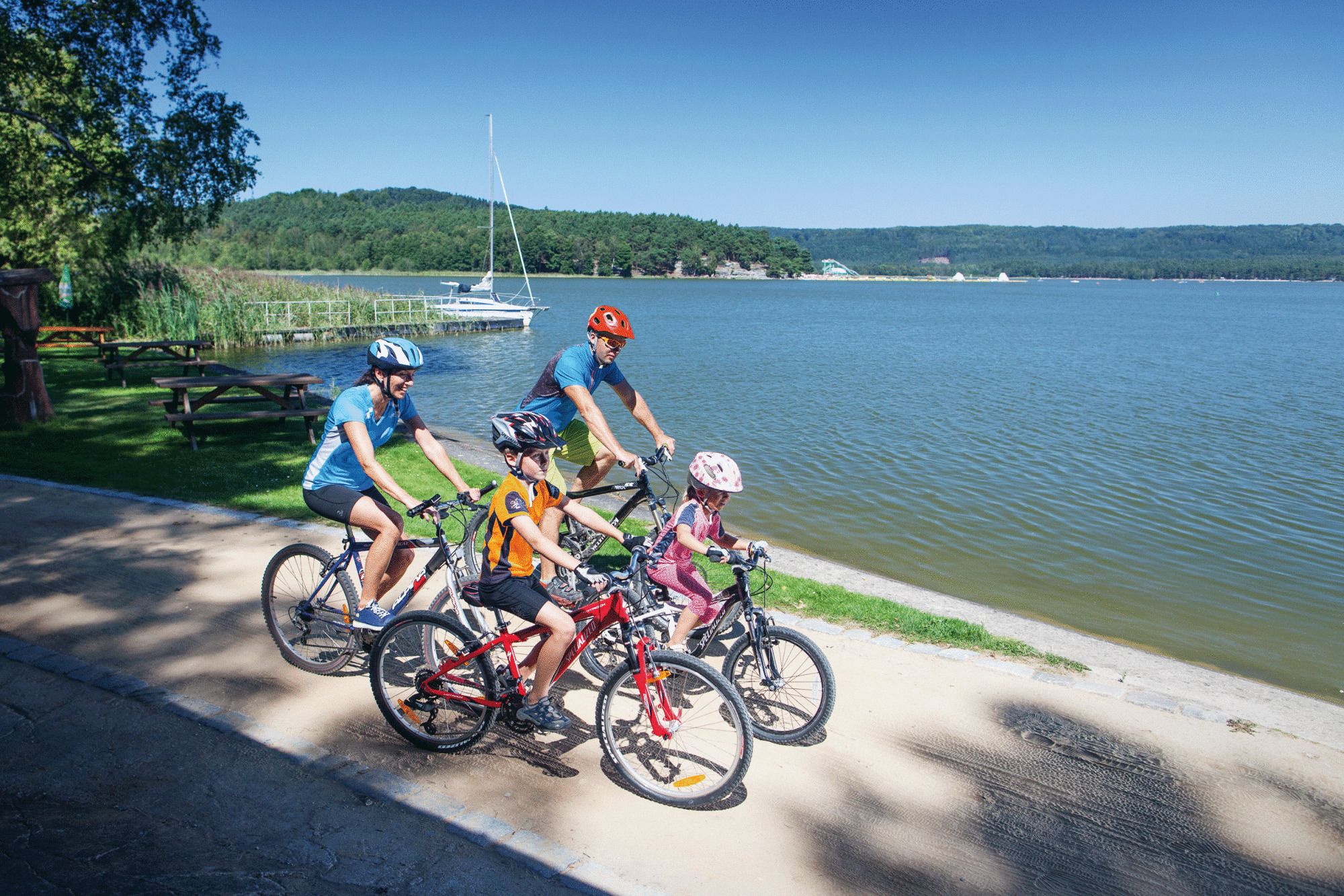 Un grupo de ciclistas en un camino pavimentado junto a un lago, con mesas de picnic y un velero al fondo.