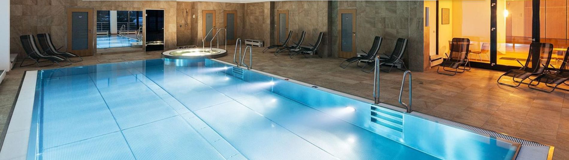 Ein Hallenbad mit klarem Wasser, Liegestühlen und Blick ins Freie.