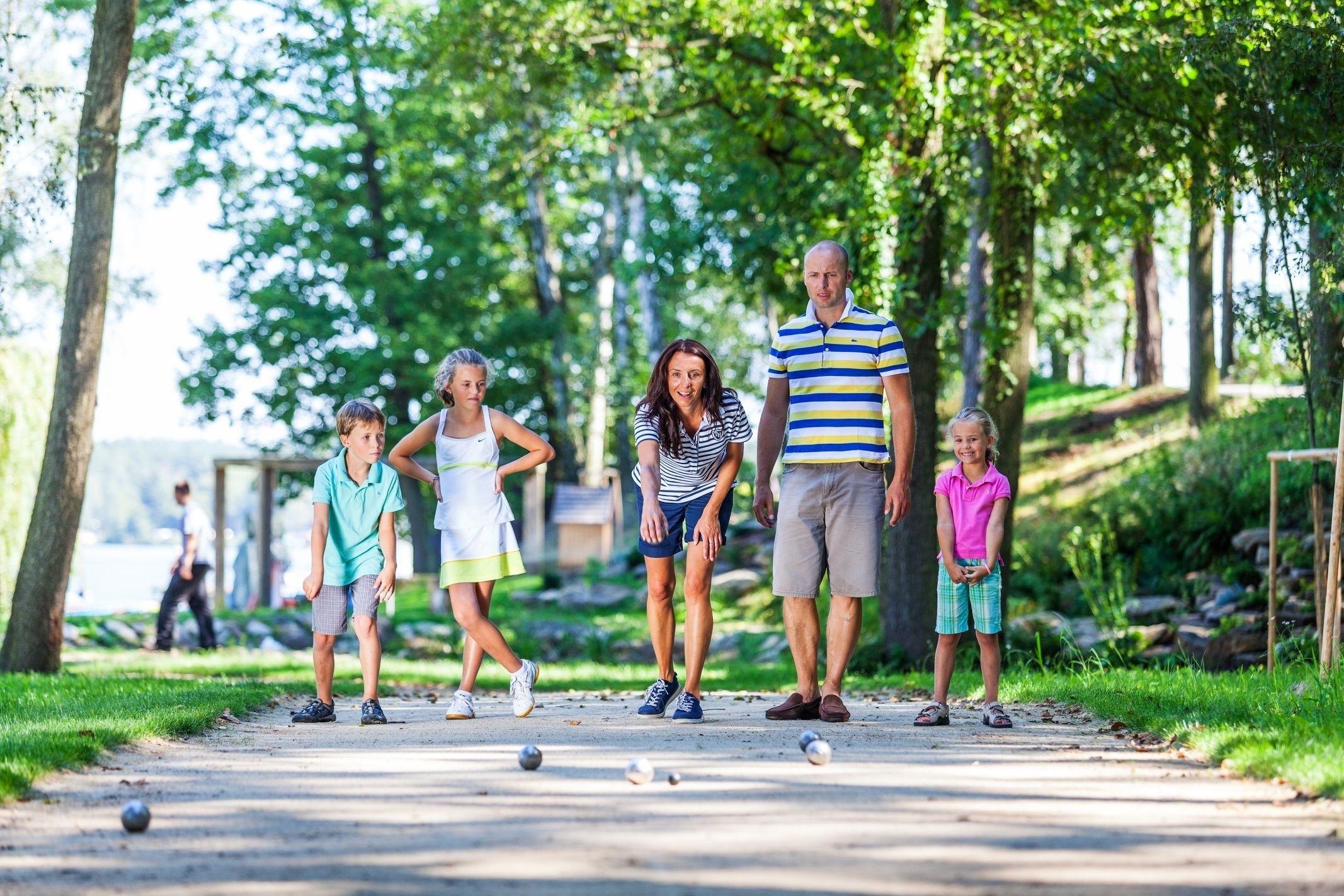A family playing croquet on a paved path in a park with trees and greenery in the background.