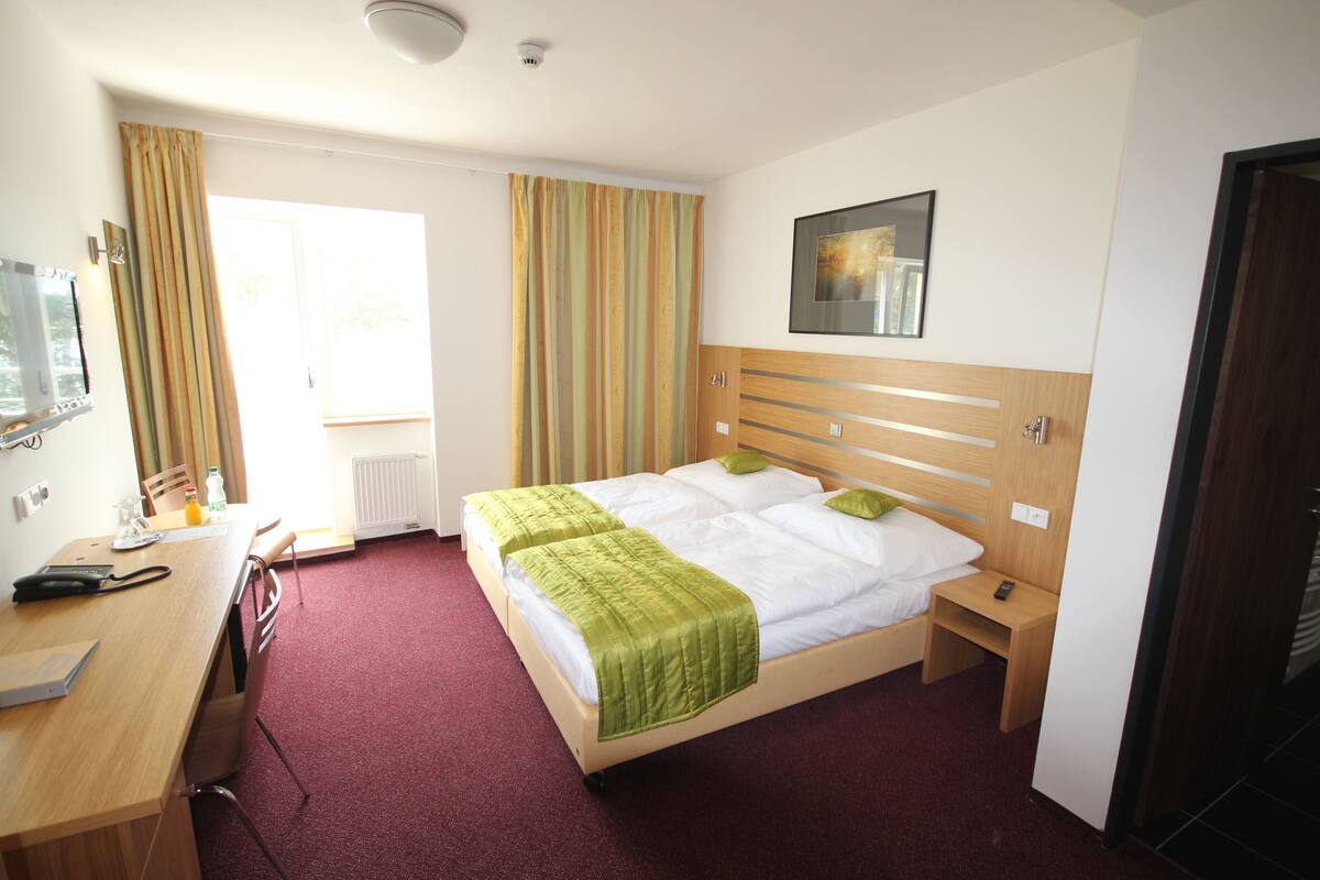 A hotel room with a bed, a desk, a chair, a nightstand, a mirror, and a window with curtains.