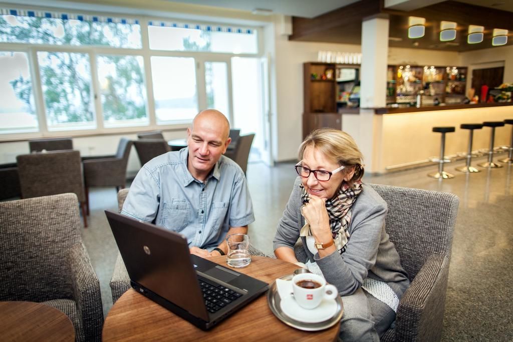 A man and a woman sitting at a table with a laptop, a glass of water, and a cup of coffee in a well-lit indoor space with a bar in the background.