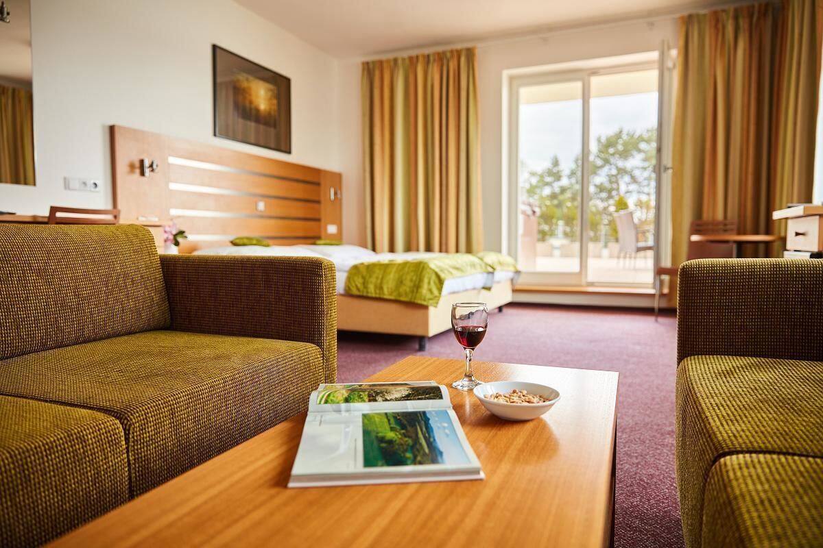 A hotel room with a bed, a sofa, a dining table with a bowl and a wine glass, and a window with curtains.