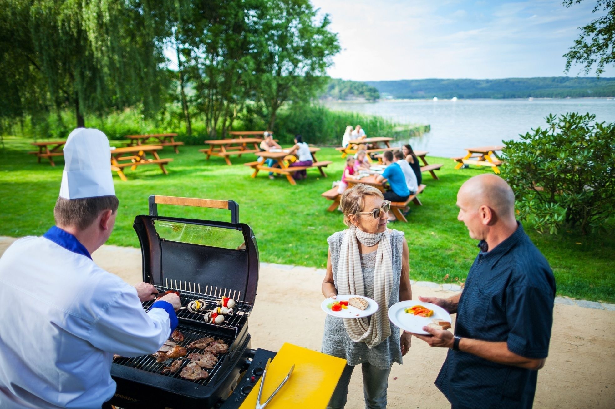 A barbecue with people around it, a lake in the background, and picnic tables.