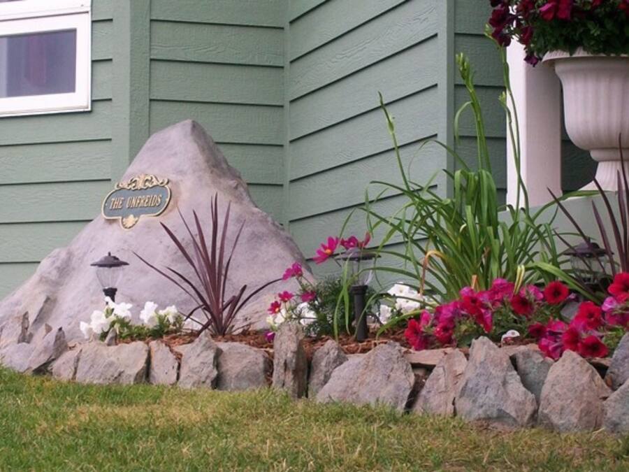 Um jardim ajardinado com uma formação rochosa, uma planta em vaso com flores rosa e uma placa com a inscrição 'The Unfreeds'.