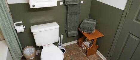 A small bathroom with a toilet, a trash can, a towel rack, and a shelf with items.