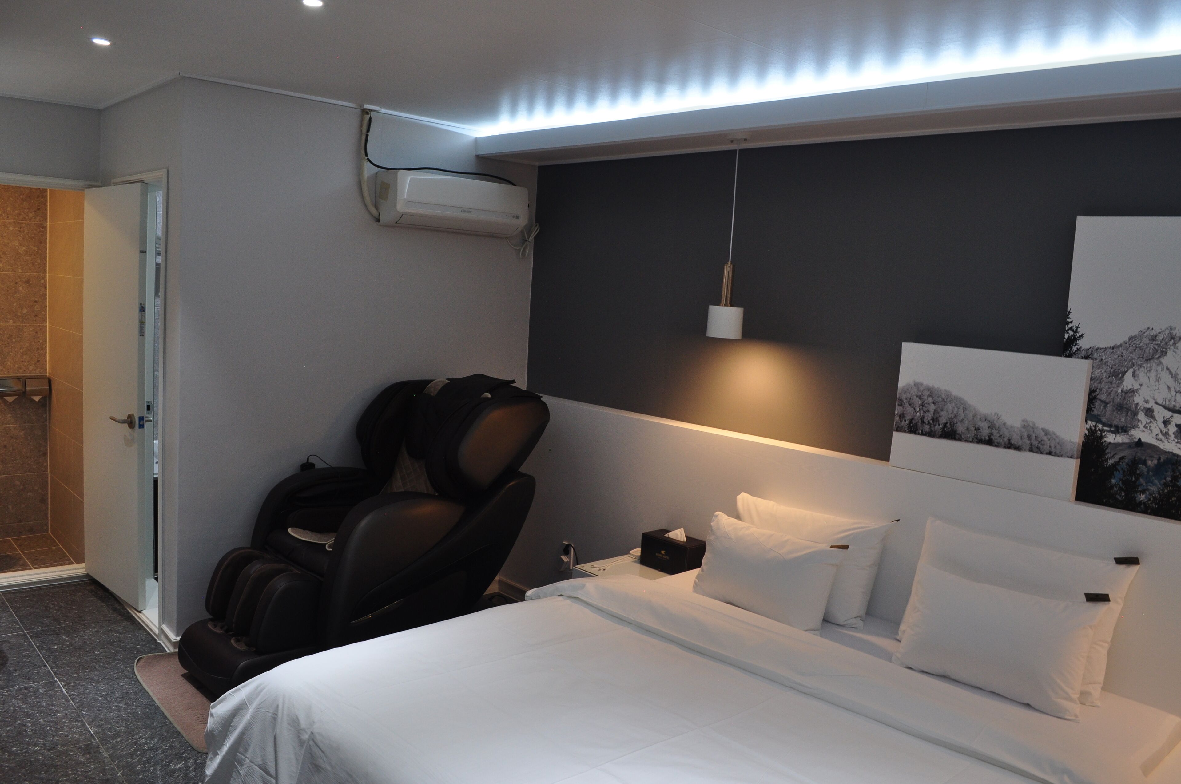 A hotel room with a bed, a massage chair, a nightstand, and a wall-mounted air conditioner.