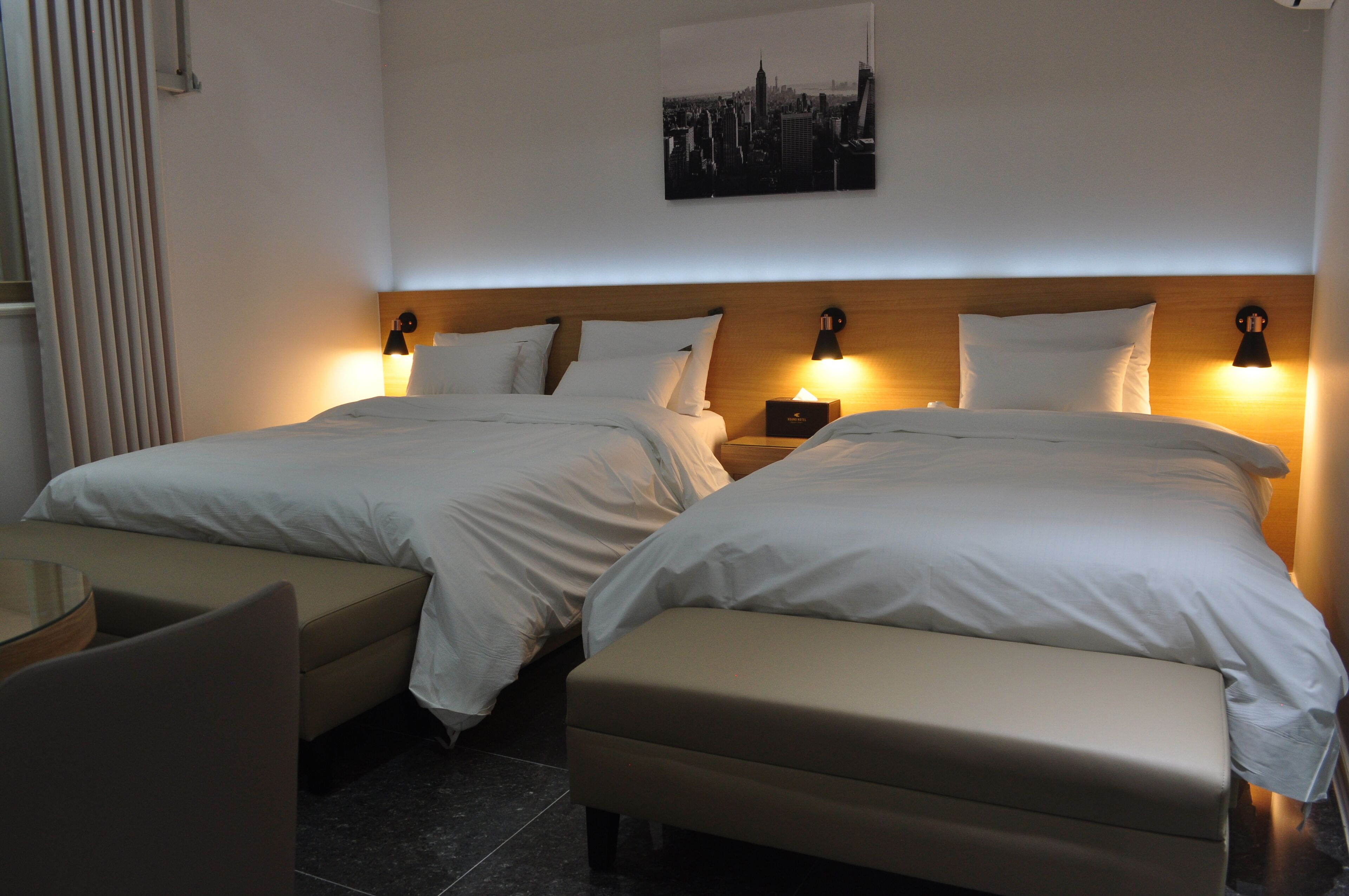 A hotel room with two beds, a small table, and a bench.
