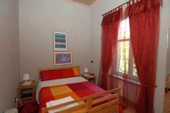 A bedroom with a bed, a nightstand, a window with red curtains, and two framed pictures on the wall. - Locanda del Barbaresco (Alba)
