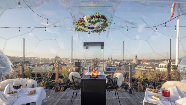 An outdoor dining area with a glass enclosure, a central fire feature, and a city view. - Pytloun Boutique Hotel Prague (Prague)