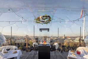 An outdoor dining area with a glass enclosure, a central fire feature, and a city view. - Pytloun Boutique Hotel Prague (Prague)