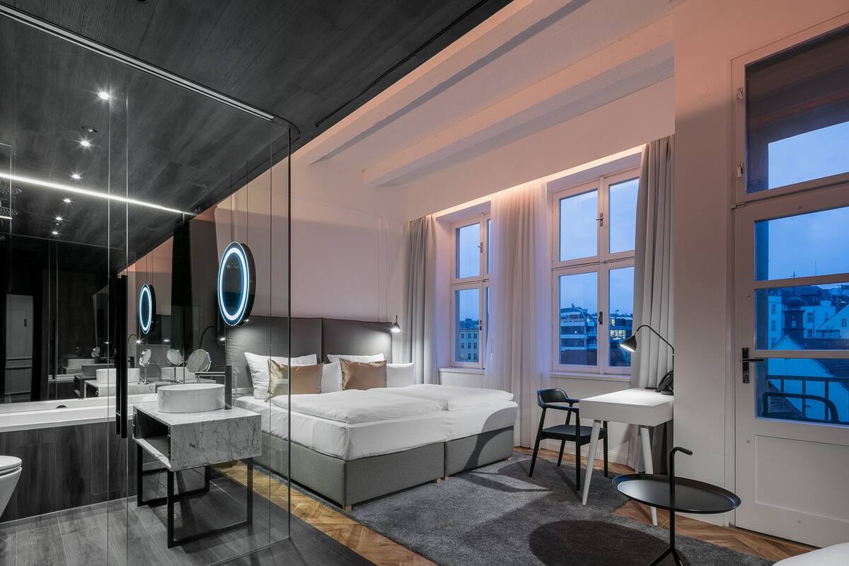 A modern hotel room with a large bed, a sitting area, a desk, and a bathroom.