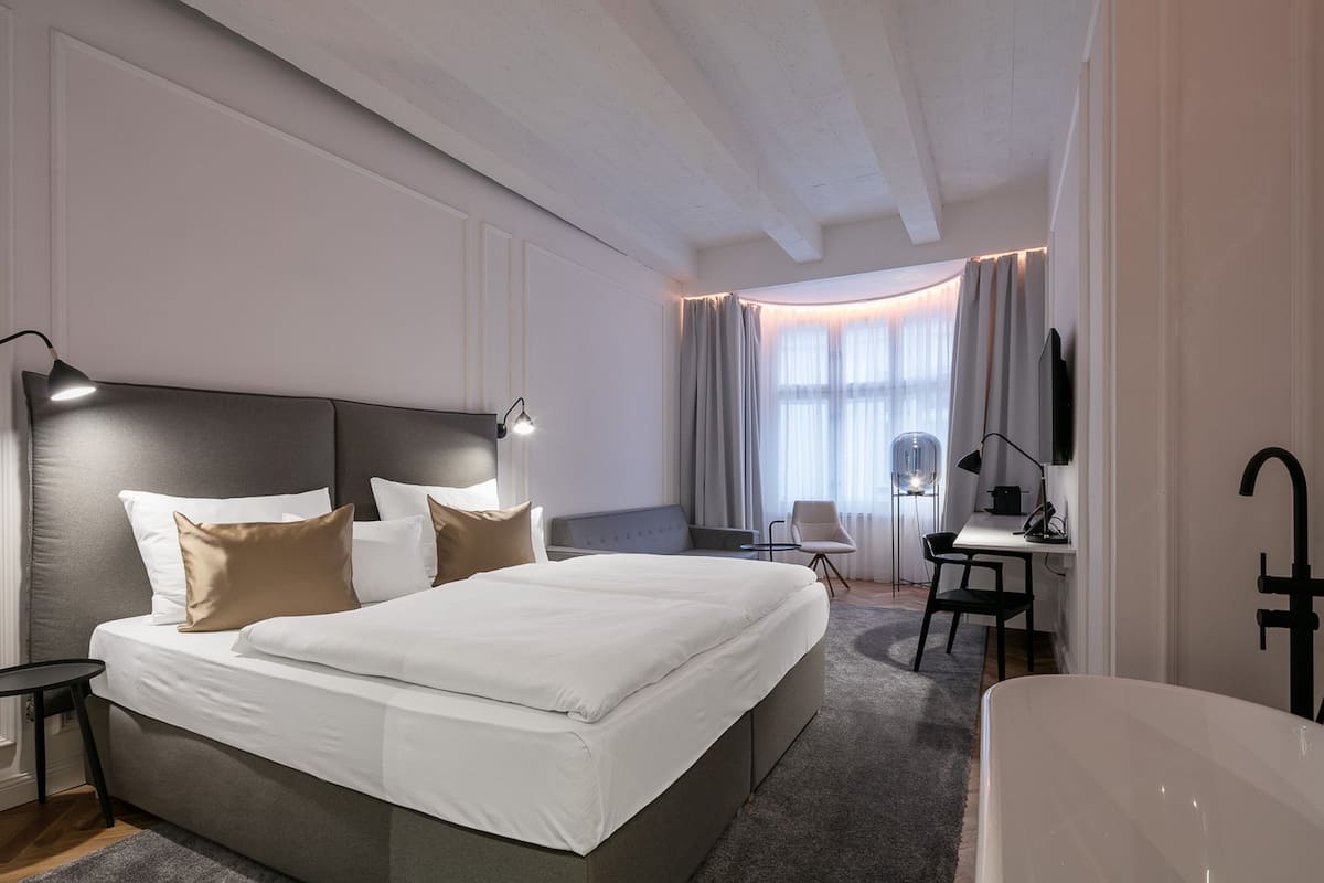 A modern hotel room with a large bed, a desk, a chair, and a bathtub.