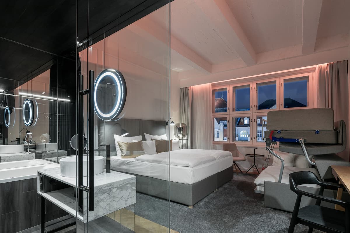 A modern hotel room with a large bed, a glass-enclosed bathroom, and a view of the city at night.