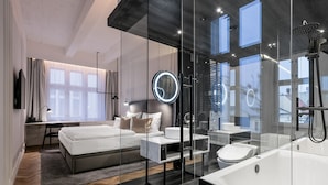 A modern bedroom with a large bed, bedside tables, a round mirror, a glass-enclosed shower, and a view of the outside. - Pytloun Boutique Hotel Prague (Prague)