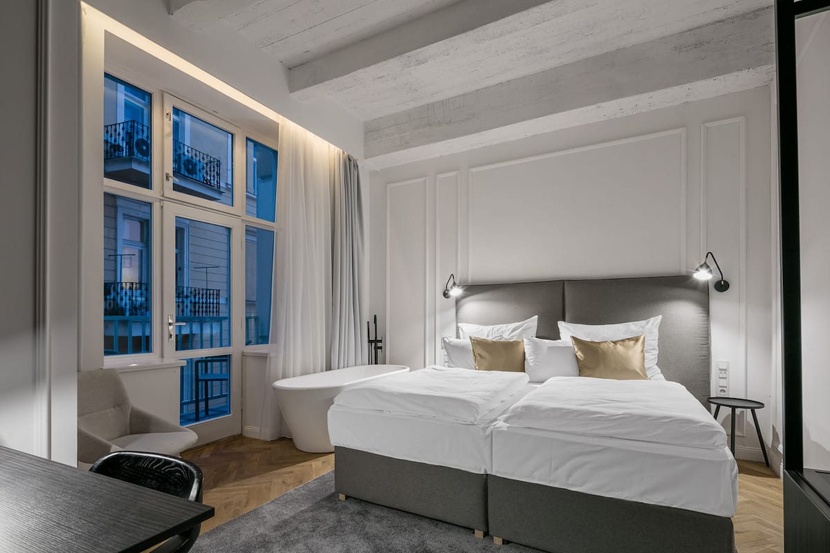A modern bedroom with a large bed, a bathtub, and a view of a blue balcony through a window.