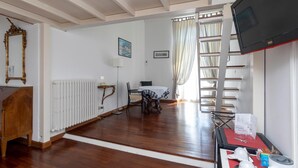 Una habitación con piso de madera, un radiador blanco, una mesa con mantel de cuadros, una silla, un televisor sobre un mueble bajo y una ventana con cortinas.