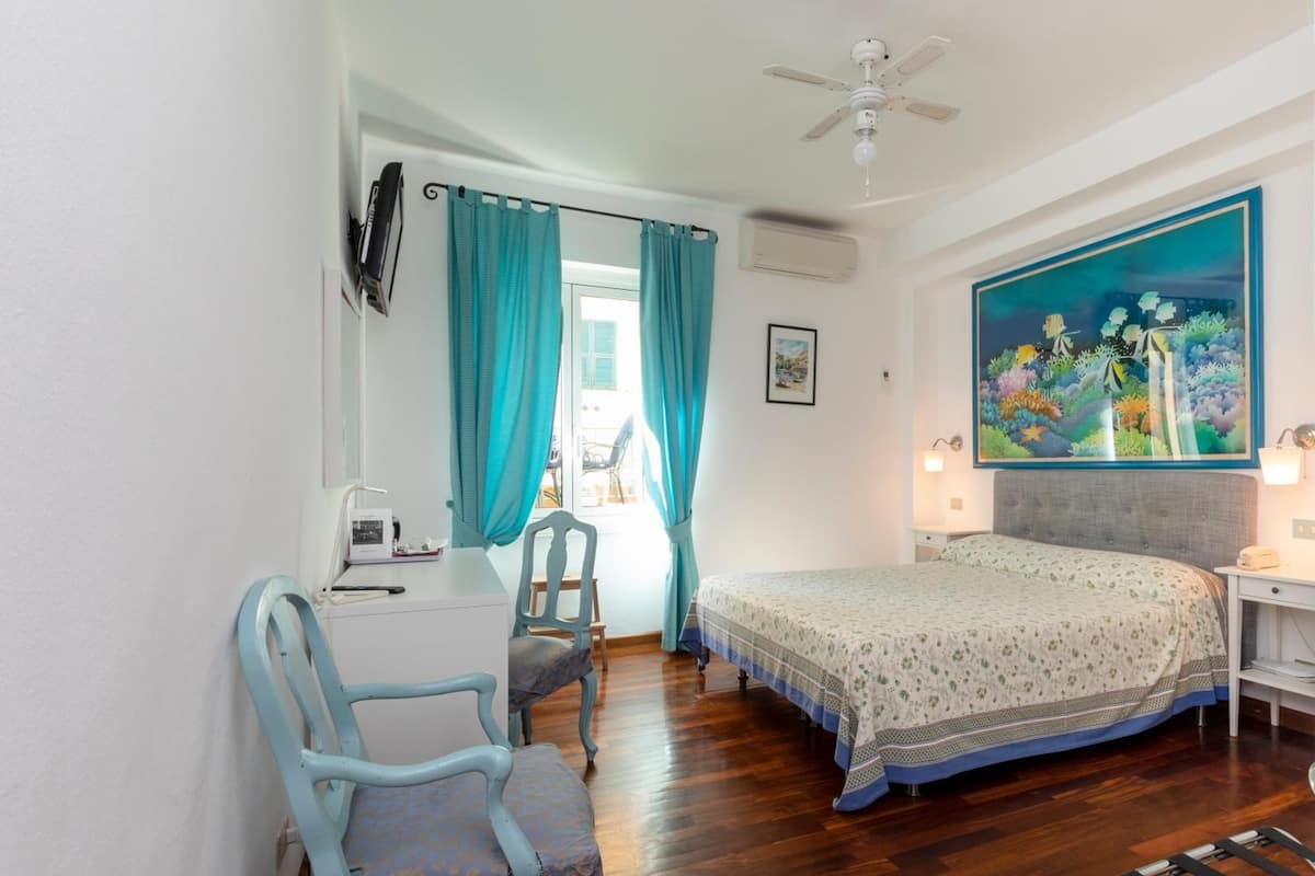 A bedroom with a bed, a desk, a chair, a ceiling fan, and a painting on the wall.