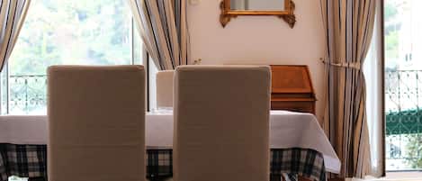 Ein Zimmer mit zwei Stühlen, einem Bett mit karierter Bettdecke, einem Nachttisch aus Holz, einem Spiegel an der Wand und einem Fenster mit Vorhängen.