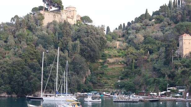 Un port de plaisance sur la côte, avec des bateaux amarrés, un château perché sur une colline et une végétation luxuriante.