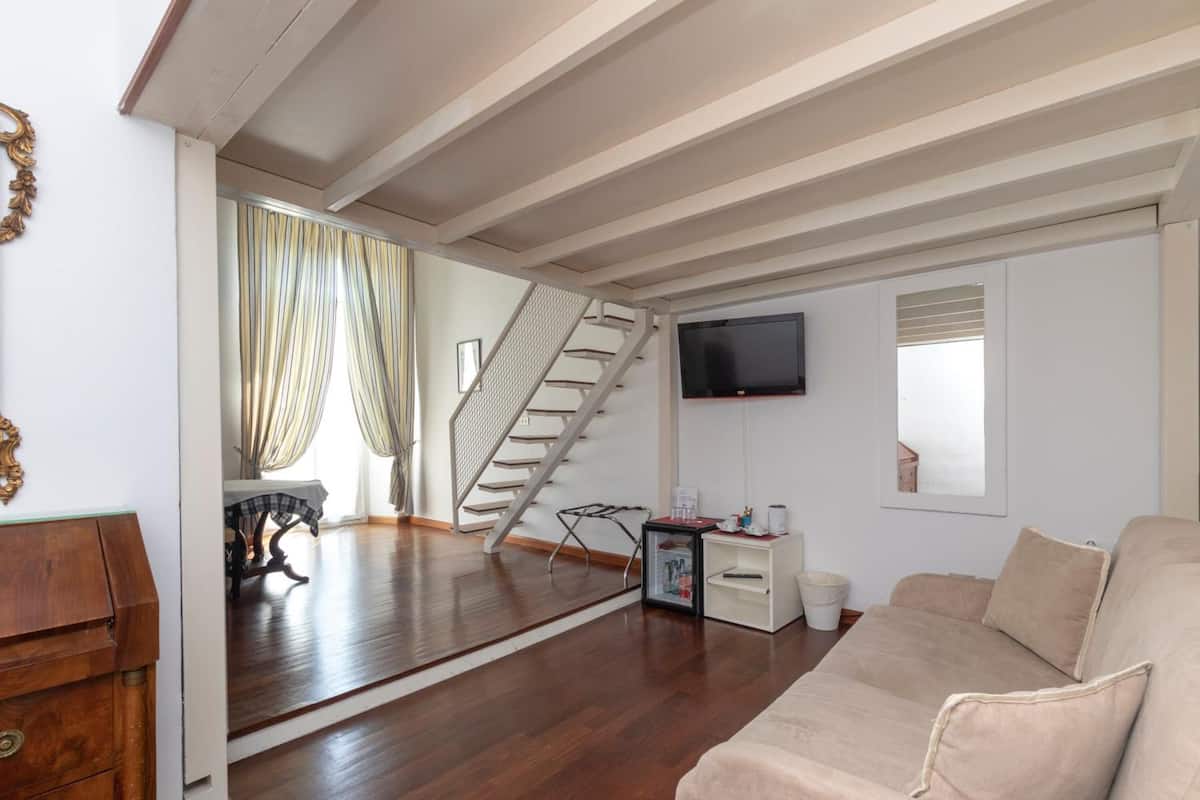 A spacious living room with a wooden floor, a beige sofa, a staircase, and a flat-screen TV mounted on the wall.