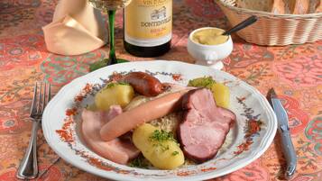 Une assiette remplie de saucisses, de jambon et de pommes de terre, une bouteille de vin et un panier de pain, le tout posé sur une nappe à motifs.