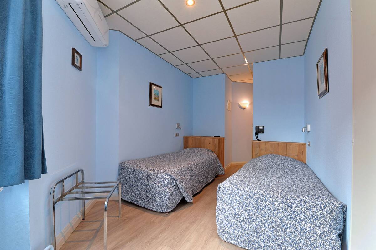 Una habitación pequeña y luminosa con dos camas individuales, cabecera de madera, radiador de metal y un calentador pequeño fijado a la pared.