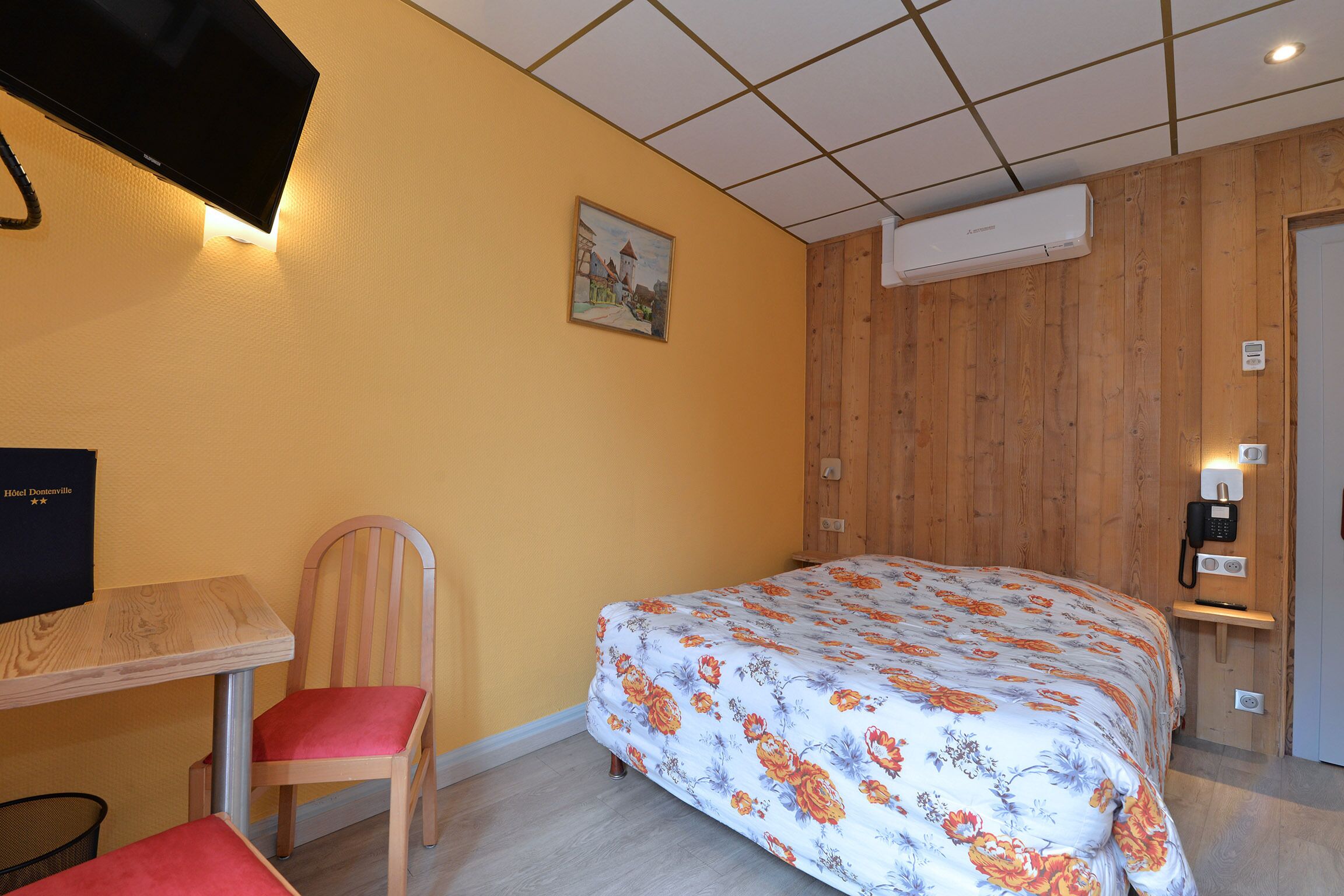 A hotel room with a wooden paneled wall, a bed with a floral bedspread, a wooden chair, a small table, and a television on the wall.