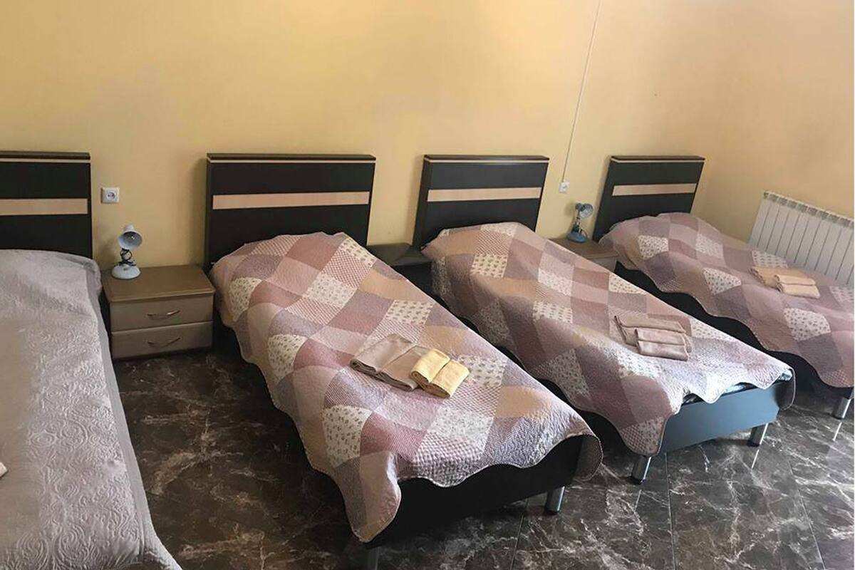 A hotel room with four single beds, each with a checkered bedspread and a pillow. There are bedside tables with lamps and a small drawer. The room has a tiled floor and a radiator on the wall.
