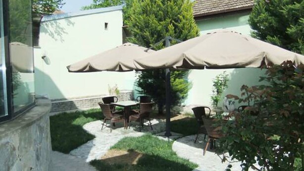 Zona de pati a l'aire lliure amb taula i cadires sota un parasol gran, envoltada de vegetació i un edifici amb teulada de rajola.