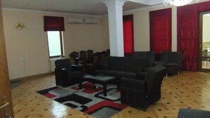 Un ampli saló amb un sofà negre gran, una taula de centre i un moqueta amb dibuixos. Hi ha dues finestres amb cortines vermelles, un penjallum i un paviment de parquet de fusta.