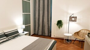 Un dormitorio con una cama, una mesita de noche con lámpara, una cortina gris, una planta pequeña, una silla de mimbre y una lámpara de pie.