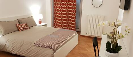 A bedroom with a bed, a bedside table with a lamp, a radiator, a TV mounted on the wall, and a potted plant.